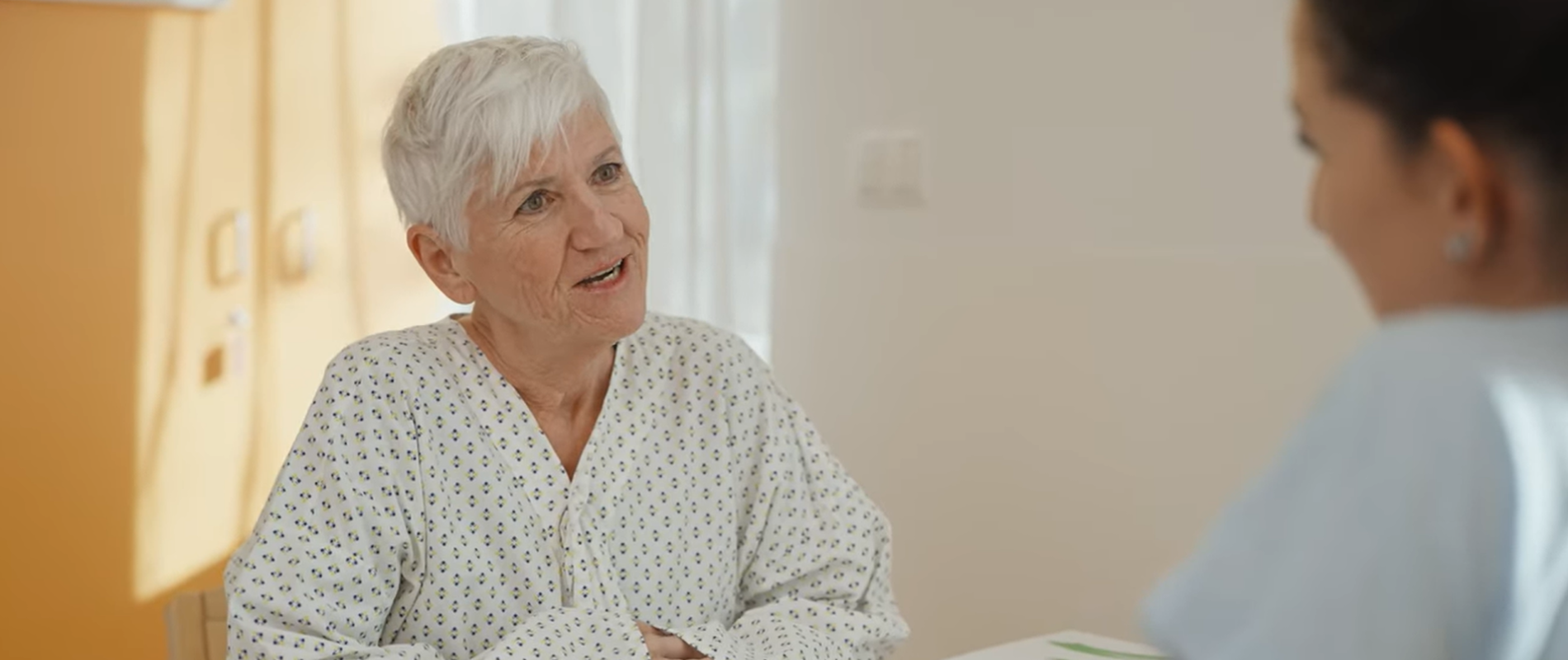 Video-Screenshot, ältere Patientin sitzt am Tisch und spricht mit Pflegerin