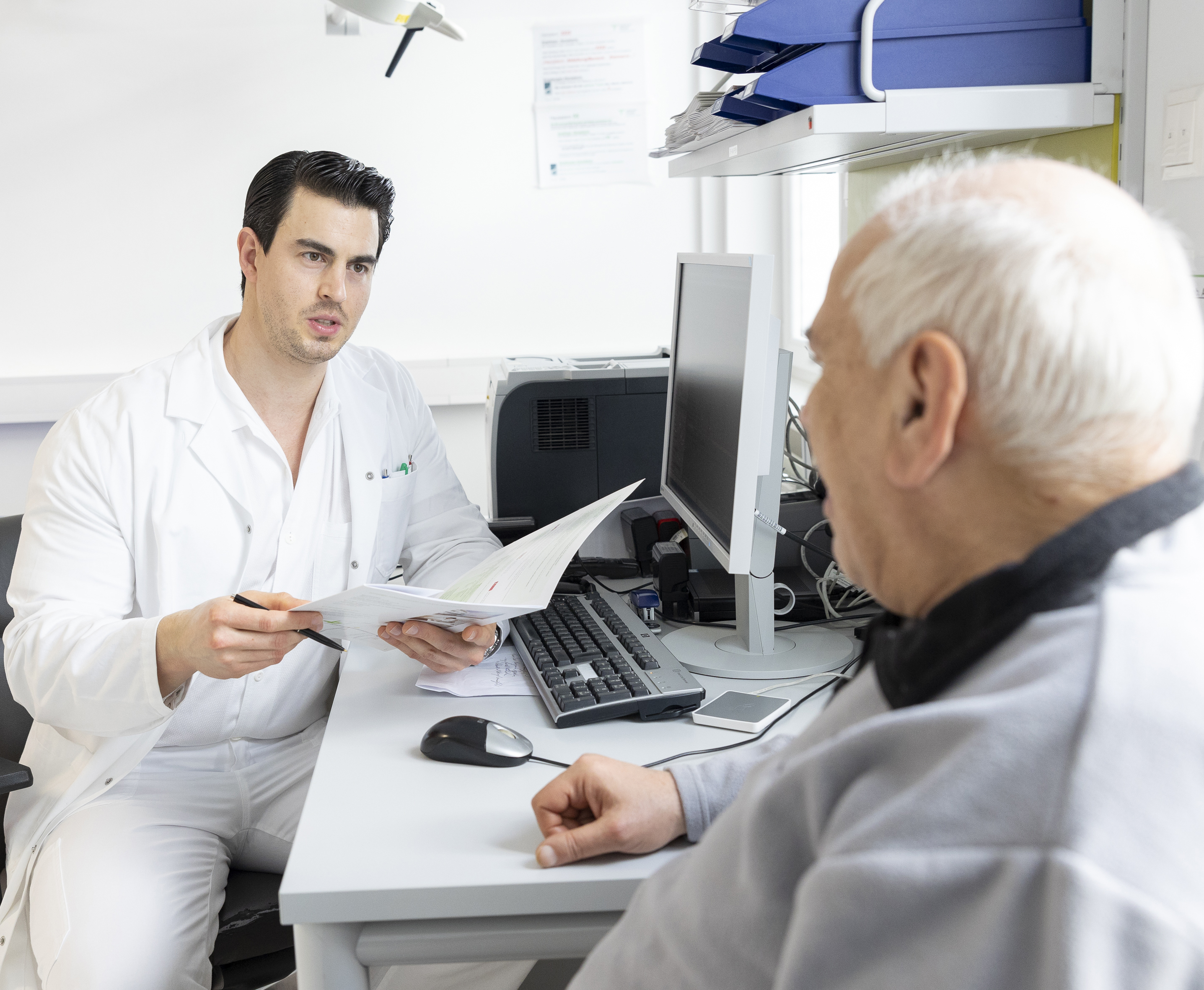 Ein Arzt im weißen Kittel bespricht Befunde mit einem älteren Patienten in einem Sprechzimmer.