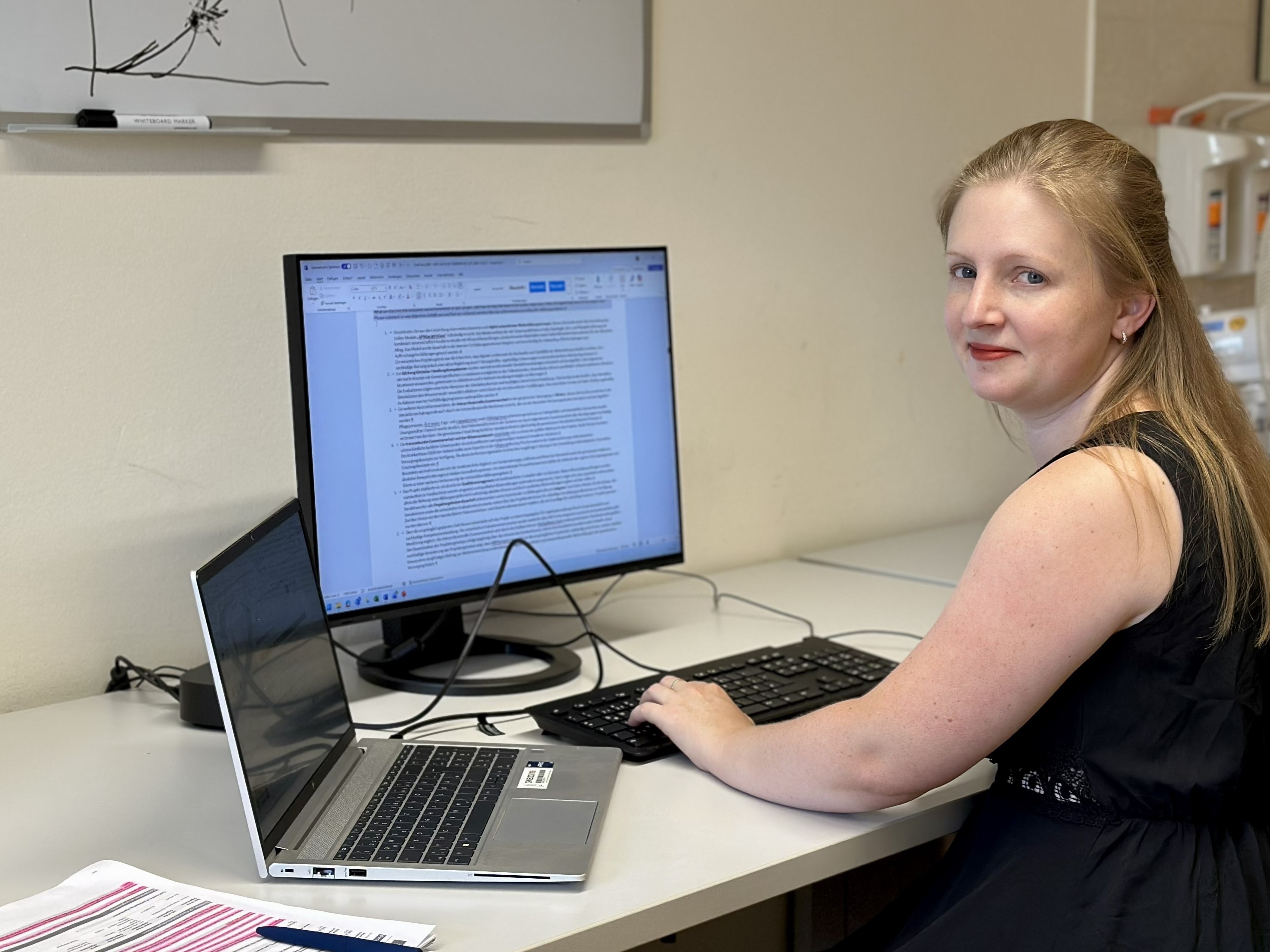Eine Frau sitzt an einem Schreibtisch und arbeitet am Computer; in der Nähe liegen Dokumente und ein Laptop. Im Hintergrund befindet sich ein Whiteboard mit Skizzen.