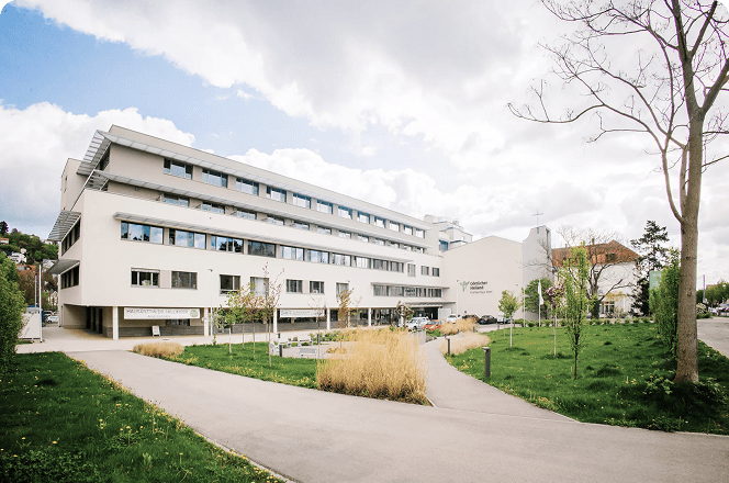 Außenansicht des Orthopädischen Spitals Speising mit einer begrünten Dachterrasse und runden Pflanzkübeln.