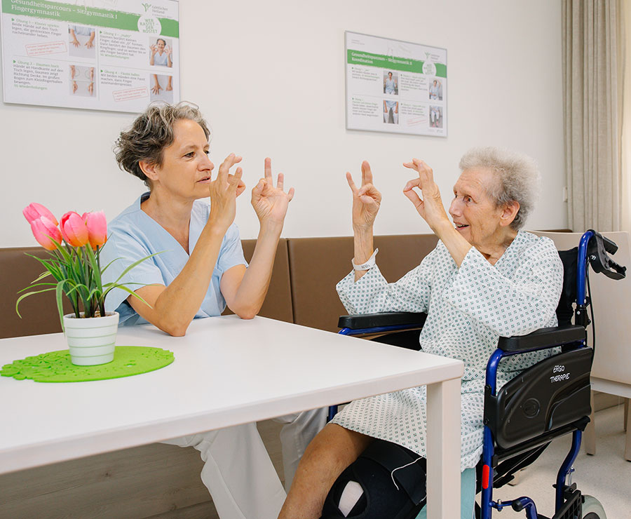 Eine Frau in Dienstkleidung sitzt neben einer älteren Patientin mit weißen Haaren und macht mit ihr Fingerübungen. 
