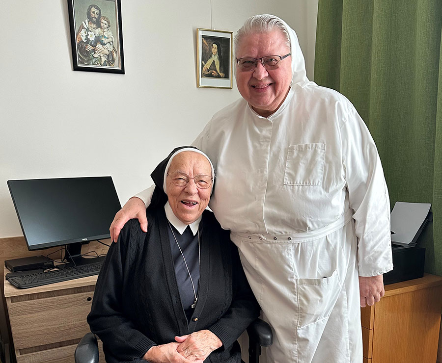 Eine Klosterschwester sitzend und eine zweite steht neben ihr und umarmt sie.